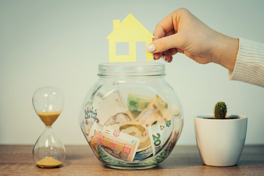Hand with symbolic paper house near glass jar with hryvnia savings. Buying new house in Ukraine concept, saving up money for a house or flat, toned photo