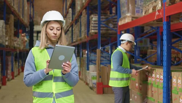 Concentrated caucasian woman using digital tablet for inventory at industry warehouse. Asian man standing behind and taking notes on clipboard while counting good on shelves.