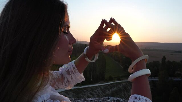 Happy woman in white boho dress making heart sign with hands on sunset in mountains. Romantic woman with long hair standing with her back on the sunset in nature in summer with open hands.
