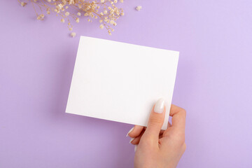 Obraz premium Female hand with white manicure holding a piece of paper with space for text. Concept of creativity, holiday wish, invitation, plans. Top view, mockup
