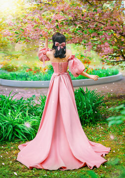 Fantasy girl princess hand touching flowers sakura tree spring garden green grass petals fall. Woman queen back rear view long pink dress puffed sleeves satin bow in hair vintage old style art photo