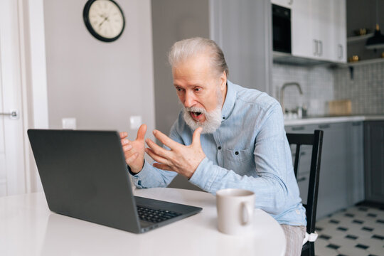Furious irritated bearded mature elderly male working from home office, angry face expression, gesturing. Angry senior older business man sitting at desk, looking at screen, having business problem.