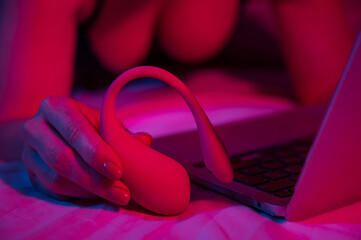 A woman is lying in bed holding a curved vibrator and looking at a laptop. Girl using sex toy in...