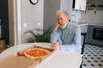 Portrait of gray-haired mature adult male eating tasty pizza thinking looking tired and bored with depression problems with crossed arms, sitting alone at table in modern kitchen room