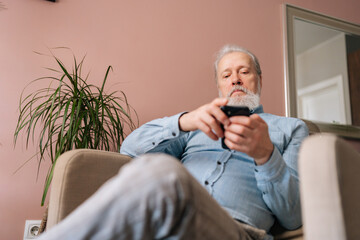 Low-angle view of serious mature male holding smartphone using mobile online app, looking to screen sitting on comfy armchair. Bearded older man enjoying social media at home after retirement.