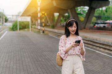 Obraz premium Asian young woman traveler with weaving basket using a mobile phone beside railway train station in Bangkok. Journey trip lifestyle, world travel explorer or Asia summer tourism concept.