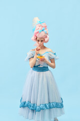 Portrait of young princess, queen wearing elegant clothes, and wig drinking cocktail over blue studio background. Vacation