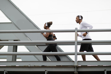 Serious young african american woman and guy in sportswear and headphones running on bridge