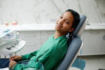 Portrait of black teenage girl sitting in dental chair and smiling at camera during checkup