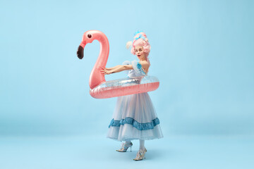 Fototapeta Portrait with funny beautiful princess, queen wearing dress holding pink inflatable flamingo and going beach over blue background obraz