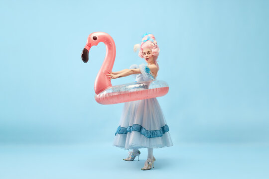 Portrait with funny beautiful princess, queen wearing dress holding pink inflatable flamingo and going beach over blue background