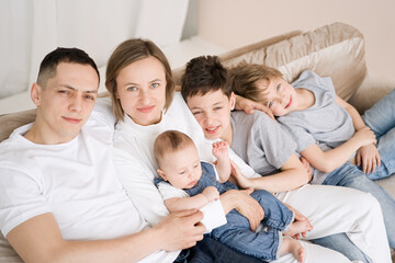 Portrait of smiling mother and father with
three sons sitting on the couch and looking at the camera, happy parents hugging children, big family posing for a photo at home