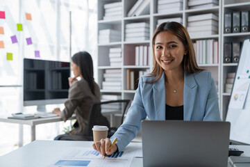 Confident asian businesswoman smiling happily relaxing holding cup of coffee drink to get rid of sleepiness and get ideas to work with laptop computer and colleagues in office.