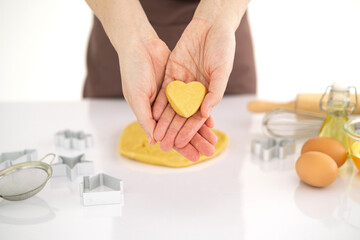 Obraz premium Women's hands offer a heart shaped cookie dough on white table background. Baking with love on Valentine's Day. anniversary. side view hands holding dough in heart shape.