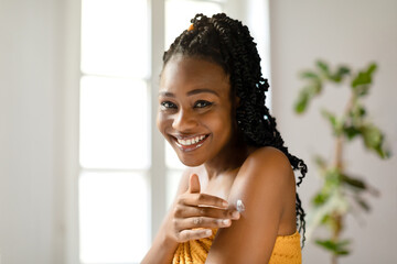 The essence of healthy skin. Happy black woman applying cream on shoulder, sitting in bedroom after shower