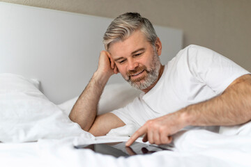 Relaxed middle aged man lying in bed, using digital pad