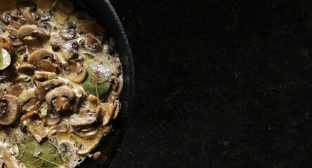 Fried mushrooms in a cream sauce with onions bay leaf and spices in a cast iron pan on a dark stone background. Top view, place for text