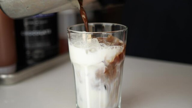 Barista stainless pitcher pours espresso and mocha sauce into glass of ice and milk, slow motion 4K