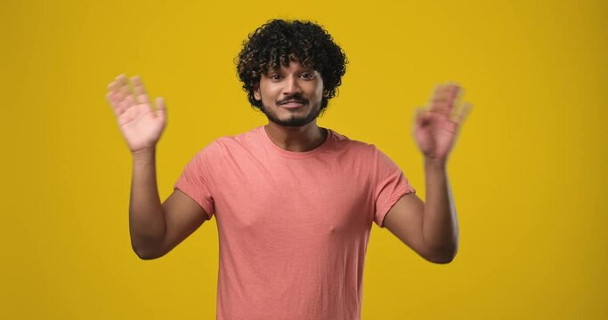 Young indian man waves hand palm in hello gesture welcomes someone expresses positive emotion standing over orange studio background