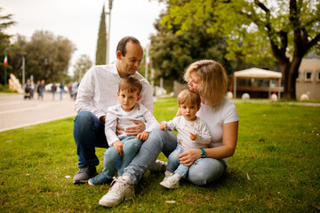 Naklejka na ściany i meble Parents and their small toddler kids son and daughter sitting on the green grass in the park and hugging each other; happy family concept