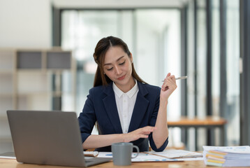 Young Asian businesswoman holding a pen sitting and analyzing the details of management information Finance is happy at the desk in the office. © crizzystudio
