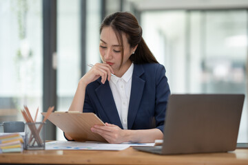 Beautiful attractive Asian businesswoman holding a clipboard of documents sitting analytically...