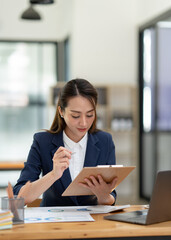 Beautiful attractive Asian businesswoman holding a clipboard of documents sitting analytically analyzing various marketing management information the finances were at her desk in the office at ease. © crizzystudio