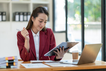 Beautiful attractive Asian businesswoman holding a clipboard of documents sitting analytically analyzing various marketing management information the finances were at her desk in the office at ease. © crizzystudio