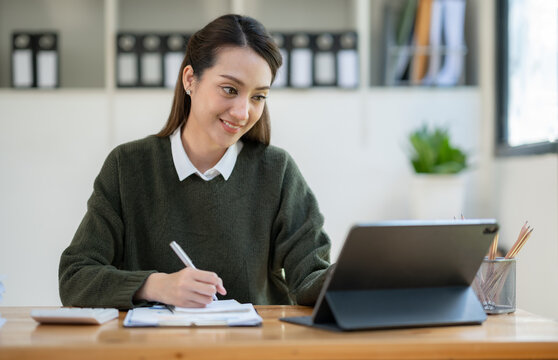 Confident Asian businesswoman happily sitting and taking notes online with a laptop computer in the office.