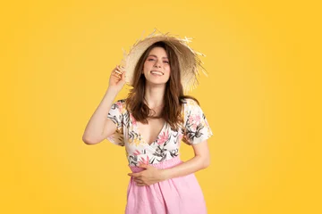 Gordijnen Pretty european lady in colorful summer outfit and straw hat, posing on yellow background and smiling at camera © Prostock-studio