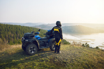  man with helmet standing near quad bike in the mountains and enjoying beautiful view of nature at sunset. - obrazy, fototapety, plakaty