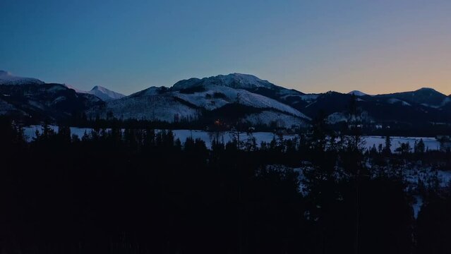 Drone Crane Shot at blue hour landing behind trees with winter snow mountains landscape