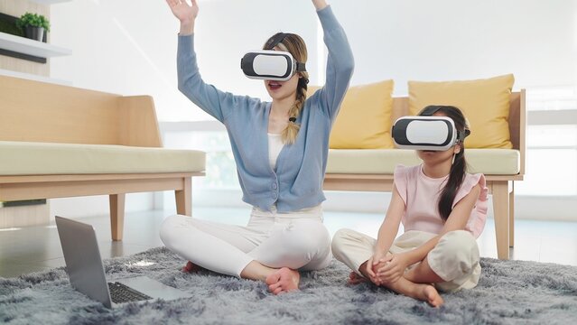 Asian mother and daughter wearing glasses of technology virtual reality headset playing game on labtop sitting on floor in living room