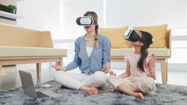 Asian mother and daughter wearing glasses of technology virtual reality headset doing yoga exercises in lotus position sitting on floor in living room. VR metaverse technology concept - Powered by Adobe