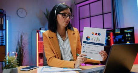 Attractive confident resolute skilled brunette in headset and glasses sitting in front of laptop during online chat and presenting report about dna facts on evening workroom background