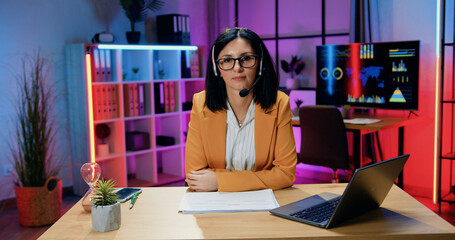 Pretty confident modern experienced brunette in headset wearing stylish clothes and eyeglasses working on laptop and looking into camera with friendly face expression in evening office
