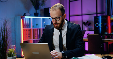 Likable confident industrious qualified bearded businessman in formal wear and in glasses sitting at his workplace and working on computer at late time in office