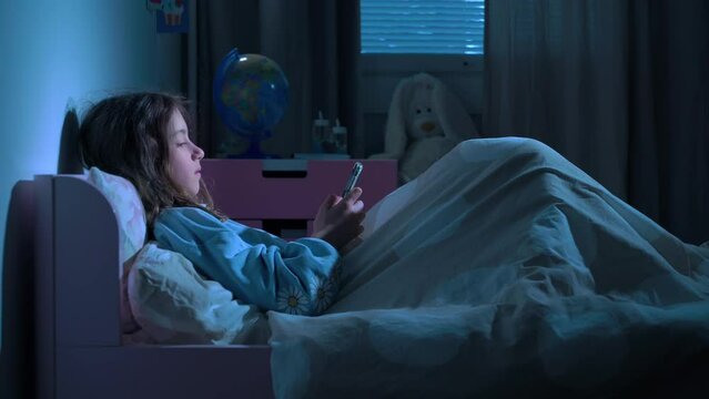 Teen girl scrolling social media texting on smartphone in bed in bedroom at night. Technology, internet, communication and people concept. Dependence on social networks in children. Not healthy sleep.