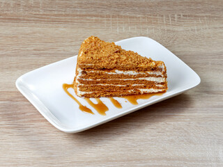 Appetizing piece of puff honey cake with cream and sprinkles, on a snow-white rectangular dessert plate standing on a wooden table.