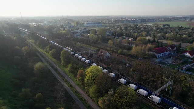 Two freight trains pass through the city in a morning drone-shot, one with empty wagons and the other transporting new delivery vans. They cross a viaduct and green terrain.