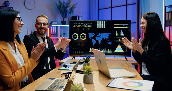 Business success concept where good-looking contented motivated experienced businesspeople discussing presentation on digital screen andclapping hands after successful collaboration