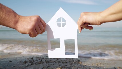 Concept of building house for family on sea coast. Dreaming of buying house by sea, family life. Hands of family hold paper house in front of sea, view from window to ocean. Symbol of home, happiness