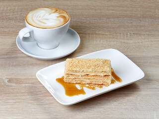 Mug of freshly brewed cappuccino with a beautiful foam is in a mug on a saucer next to a piece of Napoleon cake with honey gravy, on a rectangular dessert saucer.