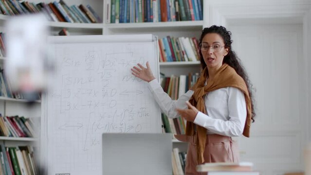 Young woman math tutor explaining topic on whiteboard to students online, streaming lesson on smartphone