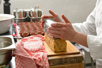 Hands of a cook toppling a savory bread pudding from a stainless steel mold onto a wooden board in a professional kitchen, selected focus