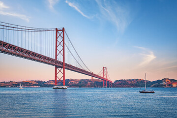 View of 25 de Abril Bridge famous tourist landmark over Tagus river, Christ the King monument and a tourist yacht boat at sunset. Lisbon, Portugal