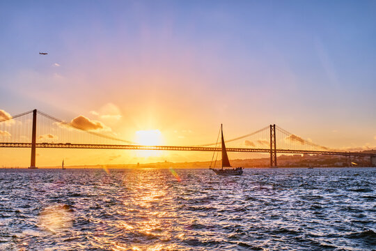View of 25 de Abril Bridge famous tourist landmark of Lisbon connects Lisboa and Almada on Setubal Peninsula over Tagus river with tourist yacht silhouette at sunset and flying plane. Lisbon, Portugal