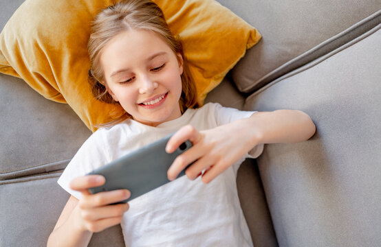Preteen girl with smartphone chatting with friends lying on sofa at home. Pretty child kid smiling with modern cellphone in social media on couch