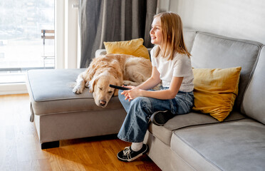 Preteen girl with golden retriever dog holding tv remote control and sitting on sofa at home....