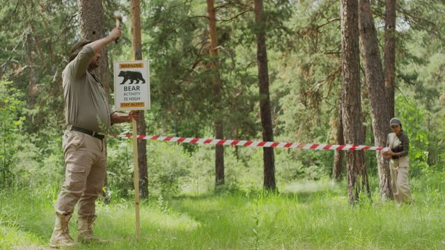Full shot of diverse male and female wardens preparing for wild animal migration season, Indian man putting up warning sign, and Hispanic woman closing off forest area with barricade tape